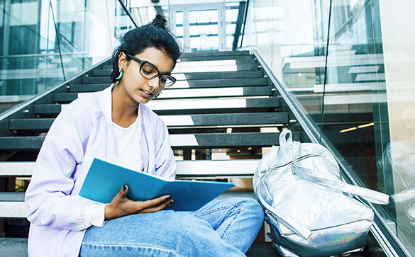 Student sitting on stairs with a note book wearing glasses